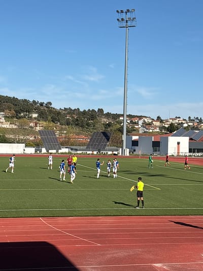 The image shows a soccer game in progress in Valpaços, Portugal. Players in blue and white uniforms are positioned on a green soccer field. A referee in a yellow shirt stands near the sideline. A red running track is adjacent to the field. A tall light pole stands near the field. The background includes buildings with solar panels and trees on a hill. The sky is blue.