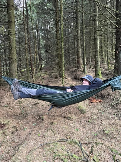 A person wearing a dark grey hoodie, black pants, and a dark grey bucket hat is lying in a dark green hammock. The hammock is strung between two trees. The person's feet are visible, wearing dark brown hiking boots. The scene takes place in a forest of coniferous trees, the ground is covered in a layer of brown pine needles. This location is within Macclesfield Forest, United Kingdom, at coordinates (latitude: 53.259, longitude: -1.991). The person is lying in a relaxed posture, gazing up at the forest canopy.