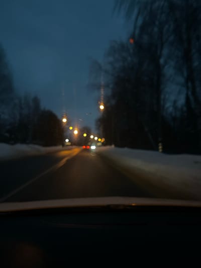 The image depicts a blurred, forward-facing view from inside a vehicle traveling on a two-lane asphalt road at dusk or night, located in Maksimkovo, Russia. Snow covers the verges on both sides of the road, which is flanked by bare trees. Multiple yellow-orange streetlights illuminate the scene, casting reflections on the wet or icy road surface. Two vehicles are visible in the distance, one with red taillights and another with white headlights. The vehicle's dashboard is visible at the bottom of the frame. No people or specific interactions are discernible. The image quality is out of focus, with some streaks or smudges visible on what appears to be the windshield.