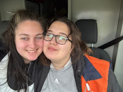 Two young adult females are depicted inside what appears to be the passenger cabin of a van or commercial vehicle. The female on the left has dark, wavy hair, blue eyes, and is smiling broadly, exposing her teeth. She wears a black and white zippered jacket. The female on the right wears black-rimmed eyeglasses and has brown, wavy hair. She is smiling subtly with closed lips and appears to be leaning her head towards the female on the left. She is dressed in a grey hooded sweatshirt with an Adidas logo, layered under a black, navy, and orange jacket. A white lanyard or cord hangs near her right shoulder. The vehicle interior features plain grey wall paneling and a dark grey fabric seat with a black headrest behind the female on the right. A black seatbelt extends diagonally across the frame. Through an opening in the upper left background, indistinct objects, possibly boxes, are faintly visible in the rear cargo area. No specific visual cues are present within the image to confirm the location as Breda, Netherlands.