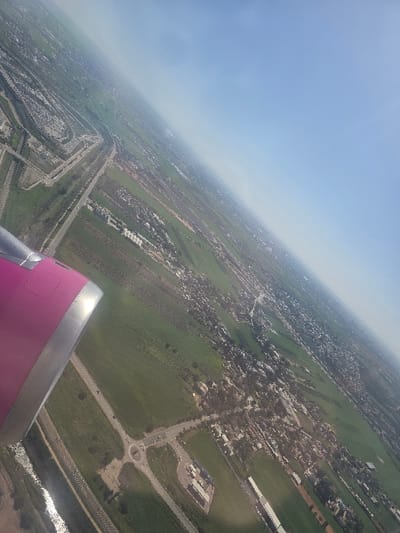 This aerial image provides a high-angle, oblique view of a cultivated and developed landscape, likely located within or immediately adjacent to the Sdot Dan Regional Council, Israel, specifically depicting areas near Ben Gurion International Airport (TLV). A portion of a magenta-colored aircraft engine nacelle with a metallic leading edge is visible in the lower-left corner, indicating the photograph was taken from an airborne airplane.

The scene is dominated by extensive green agricultural fields, segmented by roads and irrigation channels. A major multi-lane highway traverses the mid-ground, flanked by built-up areas. In the lower-middle, a large roundabout serves as a junction for several connecting roads, near which a commercial or service complex with multiple buildings is situated. A narrow, reflective body of water runs parallel to a road in the far lower-left.

Scattered residential or small community settlements, characterized by clusters of buildings and trees, are visible throughout the cultivated land. In the upper-left quadrant, a significant portion of an airport's infrastructure is discernible, featuring multiple runways or taxiways, extensive paved areas, and large industrial-type buildings, consistent with Ben Gurion International Airport. The sky in the upper-right is clear and light blue, with a slight atmospheric haze along the distant horizon. No individual persons or specific actions are discernible from this altitude.