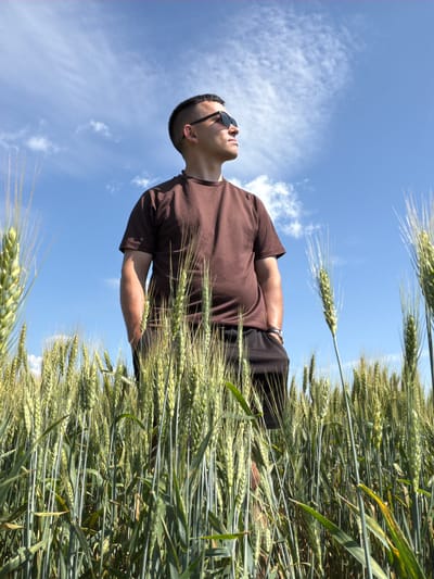 An adult male wearing a brown t-shirt, black shorts, and sunglasses stands in a field of green wheat under a blue sky with scattered clouds. He is looking up and to his right, with his hands in his pockets. The image was taken in Podshivalovo, Russia, on a sunny day.