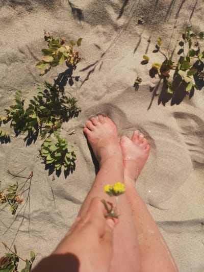 This is a high-angle, close-up shot of a person's bare feet and legs resting on a sandy surface, likely a beach given the location context of Lesnoy, Russia. The sand is light brown and shows the imprint of shadows from plants and possibly a person's body. Several small green plants with delicate leaves are scattered across the sand, some with hints of dried or reddish leaves.
The person's legs are positioned with one crossed over the other, and their feet are visible with toes pointed upwards. The skin appears smooth and tanned, with some freckles visible. In the foreground, a small yellow wildflower with green leaves is held between the person's fingers, close to their leg. The flower is out of focus, but its bright color stands out against the neutral tones of the sand and skin.
The scene suggests a moment of relaxation and connection with nature. The sunlight casting the shadows implies it's daytime, likely sunny given the distinct shadows. There is no visible text in the image. The overall mood is peaceful and serene, capturing a simple pleasure of being outdoors.