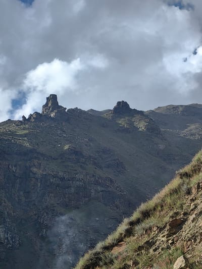 This is an exterior, medium-shot, eye-level photograph of a rocky, mountainous landscape under a cloudy sky. Two prominent rock formations rise from a ridge in the mid-ground. The left formation is tall and columnar, with jagged edges and appears to be a pinnacle. To its right, a smaller, more rounded rock formation sits lower on the ridge. The mountainside is steep and covered in sparse, dry grass and loose rocks. A series of thin, dark lines, likely power lines, stretch diagonally across the mid-ground from left to right. In the lower left quadrant, a wispy plume of white smoke rises from the mountainside. The foreground is a steep embankment covered in dry grass and rocks. The sky is filled with dramatic, gray clouds, with patches of blue visible. The image captures a rugged, natural environment, likely a remote mountainous region. No people or animals are visible. The location is identified as Yamling, India.
