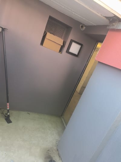 A low-angle, Dutch tilt, medium shot captures a section of a concrete floor and a grey wall. A black telescoping pole, likely part of a prop or support, stands upright on the left side of the frame, its base resting on the floor. The pole has some visible red and white stickers or labels near its base. The wall is painted a muted grey and features a square indentation with a metal grate. A cardboard box, partially opened, is placed within this indentation. To the right of the indentation, a framed picture with a white insert is mounted on the wall. Further to the right, a reddish-pink textured wall segment is visible, adjacent to a light blue wall. A reflective strip or surface is present between the grey and blue walls. The scene appears to be outdoors or in an open, unfinished structure. The image context does not provide information about the city of Hamm, Germany.