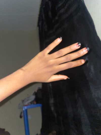 A person's left hand is extended towards the right side of the frame, with the palm facing the viewer. The skin tone is dark. The fingernails are painted with a black and red gradient design, with the black color on the tips fading into red towards the base of the nail. The hand is positioned in front of a dark, textured fabric, possibly clothing or upholstery. In the background, to the left of the hand, a blurred wall is visible. Below the hand and slightly to the left, a portion of a blue object, possibly a shelf or frame, can be seen. The lighting appears dim and artificial, creating shadows and highlights on the hand and fabric. The scene is indoors, with no specific details to pinpoint its location within Malete, Nigeria.