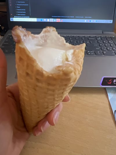 A hand is holding a partially eaten ice cream cone in front of an open laptop. The ice cream is melting and dripping from the cone. The laptop screen displays a webpage with a search bar and various icons. The background is out of focus, but appears to be a desk or table. The scene is indoors, and the lighting suggests it could be daytime.