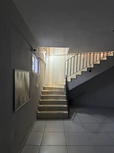 A low-angle shot looking up at a stairwell in what appears to be an apartment building in Köyceğiz, Turkey. The stairwell has concrete steps with black metal railings. Above, there is a window and a reflective object on the ceiling. The walls are white and tiled. The lighting is dim.