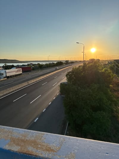 The image shows a highway with several lanes, seen from an elevated viewpoint. The sun is low on the horizon, casting a warm, golden light across the scene, indicating it is either sunrise or sunset. In the distance, there is a body of water, possibly a lake or river, and a town or village with houses. Trucks are parked on the side of the highway, and a few cars are visible on the road. There is lush green vegetation on the right side of the highway, and a row of streetlights runs along the road. The foreground shows a weathered concrete barrier, suggesting the viewpoint is from a bridge or overpass. The sky is clear with a gradient of blue and orange, typical of dawn or dusk. The overall impression is one of a peaceful, semi-rural highway scene at golden hour.