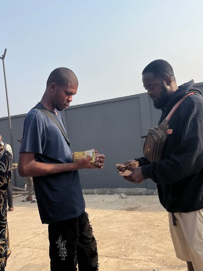 An outdoor scene in Gbonkomakent, Sierra Leone, depicts two adult males engaged in a monetary exchange or count under a clear, bright sky.

The individual on the left is a dark-skinned male with short dark hair, wearing a navy blue short-sleeved t-shirt, black distressed jeans with "AMIRI" embroidered in white on the left thigh, and a black cross-body bag strap. He holds a substantial stack of currency, secured with a rubber band, in his hands, looking down at it.

The individual on the right is a dark-skinned male with a dark beard, wearing a black hooded sweatshirt, light beige shorts, and a brown patterned cross-body fanny pack (chest bag). He holds a smaller, looser stack of currency in his hands, also looking down at the money.

In the background, a dark grey wall or building facade is visible. The ground is unpaved, light brown, and appears dry, with some debris. A dark metal pole with a V-shaped top stands on the far left. Partially visible on the far left, another person wears a black and white patterned garment. Further back, another individual's legs and feet, wearing light pants and black Adidas slides, are visible. The scene is illuminated by strong daylight.