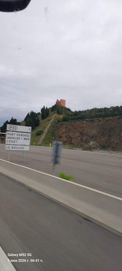 The image depicts a view from a moving vehicle on a cloudy day. In the background, a terracotta-colored building with a distinct architectural style sits atop a grassy hill. A winding pathway leads up to the building. Below the hill, a road is visible with multiple signs indicating destinations and route numbers. One sign reads "PORT-VENDRES," "ARGELÈS/MER," "CERET," and "LE BOULOU," suggesting the location is near Le Boulou, France. The foreground shows a concrete barrier alongside the road and a section of the road surface. A small, blurry green object is visible on the road near the barrier. The bottom left corner of the image displays a timestamp and device information: "Galaxy M52 5G 9 юни 2026 г. 06:41 ч."