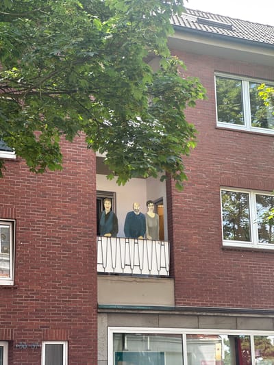 This image shows the facade of a brick building in Hamm, Germany. The building has multiple windows and a balcony area. In the balcony, there are figures that appear to be painted on or are mannequins. One figure is of a person with light skin and short hair wearing a dark blue shirt. Below that, another figure is visible, with fair skin and dark hair, also wearing a dark blue garment. They are positioned as if looking out from the balcony. The facade is made of red brick, and there are several windows with white frames. A large tree with green leaves is in the foreground, partially obscuring the building. Sunlight is filtering through the leaves, suggesting it is daytime, and the sky appears bright. The architectural style is modern. There is no visible text in the image.