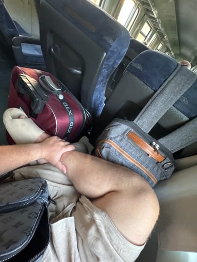 The image shows a close-up of a person's legs and luggage on a train. The person is wearing light-colored shorts and a single, light-colored slide sandal on their left foot. Their right leg is bent and resting on the seat, with their left hand placed on their right shin.
Several pieces of luggage are visible:
* A dark red duffel bag with "XTD" written in white letters on a black strap.
* A grey backpack with brown leather accents, placed behind the duffel bag.
* A black crossbody bag with a repeated grey monogram pattern, positioned in the foreground to the left.
The environment appears to be the interior of a train carriage, with blue upholstered seats visible in the background. Sunlight streams in from windows along the side of the train. The specific location of Kosovo, Bulgaria is not ascertainable from the image itself.