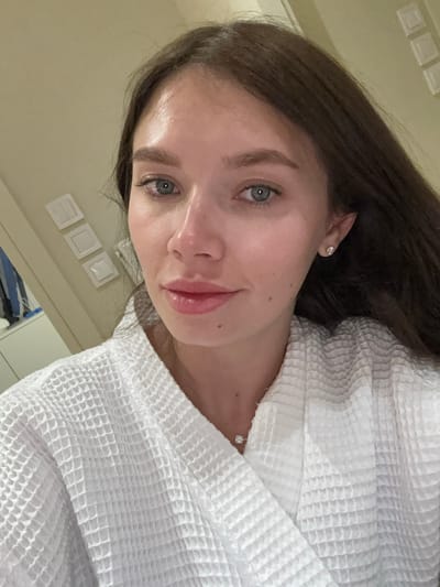 A woman is wearing a white robe and jewelry, facing the camera in an indoor setting. The location appears to be a hotel room or similar space, inferred from the wall switches and visible bedding, possibly within the Municipal Unit of Elliniko, Greece.