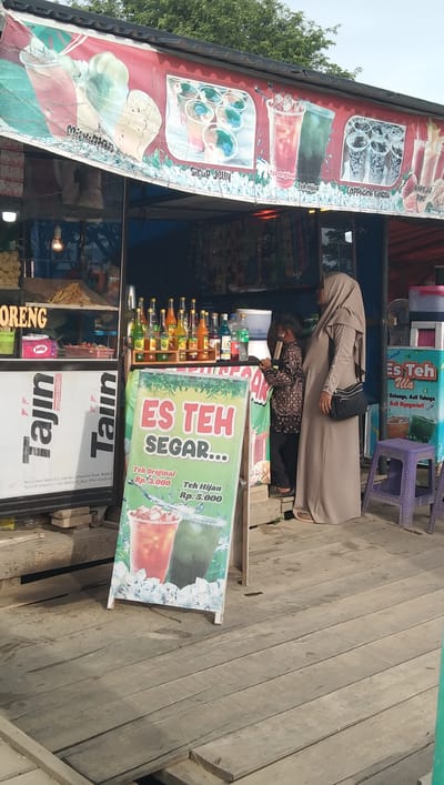 This image depicts an outdoor scene at a roadside beverage stall in Mesjid Tuha, Indonesia. The main subjects are two people: an adult wearing a long, light brown hijab and dress, and a child in a patterned shirt and pants. They appear to be interacting with the stall, with the child leaning towards it and the adult standing nearby, looking towards the stall.

The stall is set up on a wooden platform and features a prominent sign advertising "ES TEH SEGAR..." (Fresh Iced Tea), with prices listed for "Teh Original" (Original Tea) at Rp. 3.000 and "Teh Hijau" (Green Tea) at Rp. 5.000. Behind the sign, shelves are stocked with various bottled beverages and what appear to be snacks. A banner above the stall displays colorful images of drinks like "Sirup Jelly," "Teh Original," "Teh Hijau," and "Cappuccino Cincau."

Other objects visible include bottles of brightly colored syrup, a white water cooler, a purple plastic stool, and what seems to be a fan in the background. The ground is made of weathered wooden planks. The lighting suggests it is daytime, possibly late afternoon, with shadows indicating the sun's position. The overall atmosphere seems casual and typical of a local food and drink vendor. Visible text includes brand names like "Tajin" and "Jolly," along with menu items and pricing.