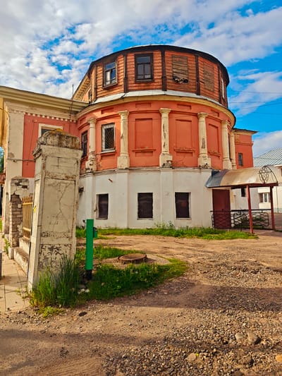 The image shows a decaying two-story building with a distinctive cylindrical addition in Faliukovo, Russia. The building's lower level is white with boarded-up windows. The upper level is a faded coral color with classical-style columns. The cylindrical addition is a darker wood at the top with multiple windows. A dilapidated concrete fence post stands in the foreground. The ground is dirt and gravel, with some grass and a manhole cover. An awning covers a doorway on the right side of the building. The sky is blue with scattered white clouds.