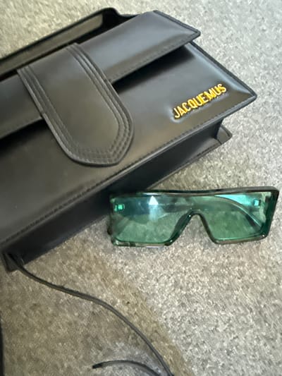 A black leather Jacquemus handbag and a pair of green tinted sunglasses are placed on a grey, textured surface. The handbag is rectangular with a flap closure and the brand name "JACQUEMUS" is embossed in gold lettering on the front. A thin black strap extends from the left side of the bag. The sunglasses have a rectangular, wrap-around style with green lenses and a dark frame. The background surface appears to be carpet or a similar textile. There are no people or animals visible in the image. The scene is static and appears to be a still life arrangement. The specific location in Hoofddorp, Netherlands, cannot be determined from the provided image.