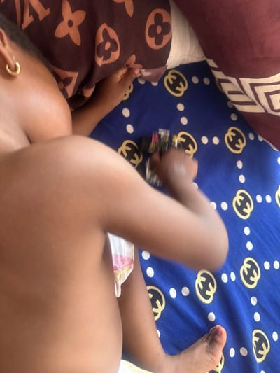 An overhead view depicts a young child, possibly a toddler, partially visible on a blue sheet with a golden interlocking "G" or "C" pattern and white polka dots, located in Iperu, Nigeria. The child's dark-skinned upper torso, right arm, and right leg are present in the frame. A single gold hoop earring is visible on the child's left ear. The child wears a white and pink patterned diaper or underwear. The right hand grasps a small, multi-colored toy, possibly a vehicle, with black and yellow details. The child's head and left side are partially obscured by a brown pillow featuring a repeated stylized quatrefoil and star pattern. Another brown pillow with a white and brown geometric pattern borders the scene on the right. The setting appears to be an indoor space, likely a bed.