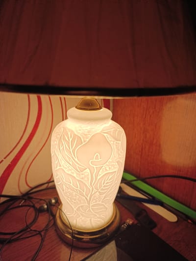 A close-up shot of a white porcelain table lamp with an embossed floral design, possibly a calla lily and leaves. The lamp is lit from within, casting a warm glow. It sits on a wooden surface, surrounded by black wires and what appear to be earbuds. In the background, a wall with red and white curved stripes is visible on the left, and a wood-paneled wall on the right. The upper portion of the image is dominated by a dark reddish-brown lampshade. The scene suggests an indoor setting, likely a home, with no discernible elements connecting it to a specific urban location within Белавушскі сельскі Савет, Belarus.