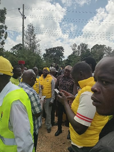 An outdoor gathering of approximately 15-20 adult males is depicted in Shirungu-Mugai ward, Kenya, on unpaved reddish-brown ground under a partially cloudy sky. The scene is dominated by several individuals wearing bright yellow padded vests, some paired with yellow caps or hard hats, indicating a potential group affiliation. On the far left, a male wears a yellow hard hat and a high-visibility yellow vest with a dark emblem. Adjacent to him, a male in a light-colored plaid shirt and glasses is observed looking down at a smartphone held in his hands. Towards the right, a male in a bright yellow padded vest over a white t-shirt with red-striped sleeves is focused on a smartphone in his hands. Another male in the mid-ground, wearing a dark patterned shirt and a light-colored cap, also engages with a smartphone. In the background, a wooden utility pole with multiple cross-arms and numerous overhead lines is visible on the left, set against a backdrop of dense green foliage and trees. No specific interactions beyond general gathering and smartphone use are clearly discernible.
