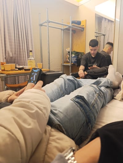 The scene is an interior of a room, possibly in a hotel or apartment in Bobigny, France. Two people are present. One person is lying on a bed, facing towards the camera, wearing blue jeans, a beige jacket, and holding a small device in their hands. The screen of the device is visible, displaying a figure. The other person is sitting on the bed behind, wearing a dark t-shirt and dark pants. They are also holding a phone and looking at it.
The room has a casual, lived-in appearance. There is a table with a bottle of Jack Daniel's whiskey, some cups, and food items on it. Behind the table is a luggage. A metal shelf structure is visible in the background, holding items like a portable speaker and a grocery bag. A mirror is on the wall, reflecting the seated person and the room's interior. The curtains are partially drawn, letting in light.