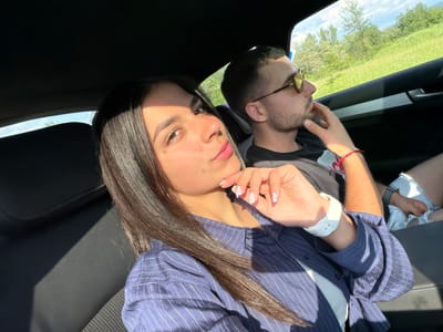 A couple is in a car. The woman in the foreground is wearing a blue striped shirt and a white watch, holding her hand near her face. The man in the background wears sunglasses and a black T-shirt, with a hand near his chin. They appear to be driving through a green, rural area. Given the location provided, the image may have been taken near Хаджийско, Bulgaria. The interior of the car is dark, with leather seats.