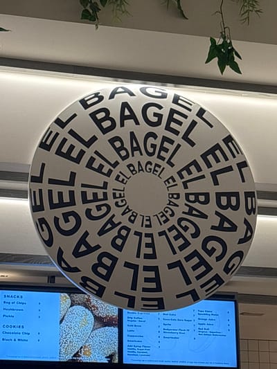The interior of an "El Bagel" establishment in Winchester, United States of America, is depicted from a low-angle perspective, focusing on an overhead circular sign and digital menu boards below. The prominent, central object is a large, white, round sign suspended from the ceiling. The sign features the words "BAGEL" and "EL BAGEL" printed in black uppercase letters, arranged in concentric circles that spiral inward towards the center. Above the sign, a white ceiling with a subtle gray trim is visible, from which decorative artificial green foliage hangs in various spots. Below the sign, two illuminated digital menu screens are partially visible. The left screen lists "SNACKS" including "Bag of Chips," "Hashbrown," and "Pickle," and "COOKIES" such as "Chocolate Chip" and "Black & White." A close-up image of a sesame seed-covered bagel or pastry is displayed on this screen. The right screen details various "Double Espresso," "Drip Coffee," "Cold Brew," "Latte," "Cappuccino," and "Americano" beverages, along with "Add Syrup Flavor" options including "Vanilla," "Caramel," "Hazelnut," and "Lavender." Further beverage options listed include "Diet Coke," "Coca-Cola Zero Sugar," "Sprite," "Bodyarmor Flash IV," "Strawberry Kiwi," "Smartwater," "Topo Chico Sparkling Water," "Orange Juice," "Apple Juice," and multiple "Red Bull" varieties. No individuals are present in the scene, and no specific actions or interactions are occurring. The environment appears clean and well-lit by artificial light sources.