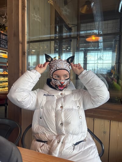 An individual is seated at a wooden table inside a commercial establishment in Vasil'evo, Russia, consistent with a cafe or restaurant environment. The individual wears a glossy white puffer-style winter jacket, bearing the "BOSCO" logo on the left chest. A unique balaclava covers the individual's head and lower face. This balaclava features a leopard print design on the upper head with two prominent dark, pointed ears. The lower face section of the balaclava displays a realistic graphic print of an open-mouthed, roaring tiger. The individual's eyes are visible through the balaclava, directed slightly upwards and to the right. Both hands are raised, with fingers touching and adjusting the ears of the balaclava. The background consists primarily of large glass windows with dark wooden frames, providing a view of a bright outdoor scene. Reflections on the glass show interior elements, including warm-toned pendant light fixtures and decorative string lights. To the immediate left, a display case stocked with pastries and bottled beverages is partially visible, along with a menu board displaying Russian text, specifically "БУРГЕР" (Burger) priced at 680 Rubles. A dark chair back is visible behind the individual, and a partial view of another person's arm/shoulder is present in the lower-left foreground.
