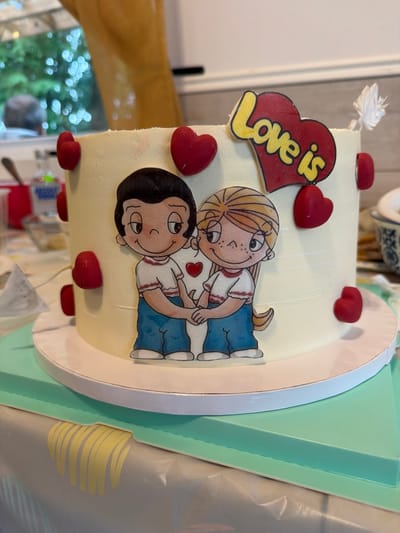 A close-up, eye-level shot shows a two-tiered round cake decorated with fondant and edible images. The cake is primarily covered in a pale yellow frosting. On the front of the cake, two cartoon characters, a boy and a girl holding hands, are depicted. Above them, a red heart-shaped decoration with "Love is" written in yellow is placed. Small red heart-shaped candies are affixed to the sides of the cake. The cake sits on a white cake board, which is then placed on a light green square base. The background is blurred, showing a window with greenery and bokeh lights outside, suggesting an indoor setting with natural light. A bottle of Absolut vodka and other kitchen items are faintly visible in the background to the left. The overall scene appears to be a domestic environment, possibly a home kitchen or dining area in Saint-Césaire, Canada.