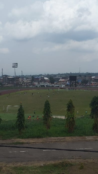 The image shows a soccer field in Buea, Cameroon, located at latitude 4.163 and longitude 9.279. The field is green, with a soccer goal at one end. There are several people on the field, some of whom are playing soccer, and others who are sitting and watching the game. The game appears to be in progress, with the players moving across the field. There are trees surrounding the field, and a paved road in the foreground. Behind the soccer field are houses, and a large billboard. The sky is overcast with white clouds.