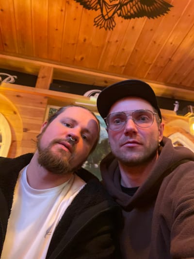 Two fair-skinned men pose in a selfie. One wears a black cap and wire-rimmed glasses with a brown hoodie. The other, with a short brown beard, has a nose ring and wears a white t-shirt and a black fleece jacket. The background includes light brown wooden walls, a decorative black eagle design, and interior lighting. The photo is oriented vertically, rotated slightly counter-clockwise.