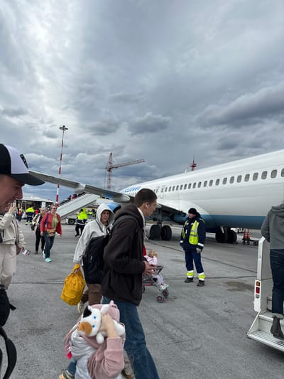 A daytime scene unfolds on an airport tarmac, showing numerous passengers queuing to board a large white and blue passenger aircraft under a heavily overcast, cloudy sky.
In the foreground, several individuals are partially visible, including one person wearing a black and white baseball cap. Further back, a line of passengers waits to ascend the mobile passenger stairs leading into the aircraft. Among them, a person in a dark jacket carries a yellow bag, and another holds a white and brown stuffed animal. A baby stroller containing a doll is also present on the tarmac.
The airplane dominates the midground, its white fuselage featuring rows of windows and a blue underside. The partial text "UTair" is visible on its side near the front. Airport ground staff, dressed in high-visibility yellow and blue jackets and dark pants, are positioned near the aircraft's landing gear and the base of the boarding stairs, overseeing operations.
The tarmac is dark grey asphalt. In the background, a large red and white construction crane and a tall light pole with aviation markings are visible against the gloomy sky. The weather appears cool and cloudy, characteristic of an overcast day. The overall activity depicted is the routine process of boarding an aircraft. The scene is located in Bolshoye Savino, Russia.