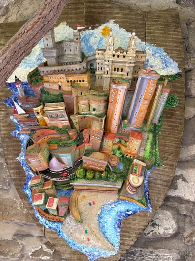 The image is a detailed, three-dimensional diorama depicting a stylized coastal city, possibly inspired by Dolceacqua, Italy, or a similar Mediterranean town. The diorama is mounted on a rustic, wooden board that resembles aged planks, framed by a rough, bark-like material on the left and stone on the right.
The city itself is a vibrant, colorful assemblage of buildings, structures, and natural elements, all rendered with thick, textured paint that gives the impression of a miniature world. In the upper left portion of the diorama, a large, grey fortress-like structure with turrets and battlements dominates the scene. A small, white sailboat with a red stripe is visible in the blue water to the left of the fortress. Adjacent to the fortress, a lighthouse stands tall.
Further into the city, a grand, palatial building with twin towers and arched windows is depicted. A bright yellow sun with radiating rays is painted in the sky behind this structure. Various other buildings of different shapes and sizes, some cylindrical and tall with visible window details, are clustered together. The architecture suggests a blend of historical and more modern elements. Buildings are painted in warm earthy tones, oranges, yellows, and muted reds, with green foliage interspersed throughout. Roads, painted in black and red, wind through the city, and a red sports car is shown on one of these roads.
The diorama's base appears to be a rough, wavy representation of the coastline, with blue paint simulating water. A sandy area with small, colorful flags is also present near the water's edge. The overall impression is of a bustling, picturesque coastal town, presented in a highly artistic and somewhat whimsical style. There are no visible people or discernible actions occurring within the diorama. The focus is entirely on the architectural and environmental representation of the miniature city.