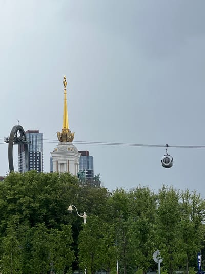 The media file depicts an outdoor scene in Vёшки, Russia, characterized by a bright, overcast sky, suggesting daytime. The primary subjects are architectural elements and natural scenery. Dominating the central portion of the frame is a tall, ornate golden spire, possibly part of a church or monument, which is juxtaposed against modern glass-fronted buildings. In the foreground and right side of the image, lush green trees provide a natural backdrop.
An interesting object suspended from wires, likely part of a cable car system or similar transport, is visible in the upper part of the frame. This object has a rounded, metallic appearance with visible panels. A street lamp with a distinctive curved arm and a glass-encased light fixture is also present amidst the foliage.
The overall scene appears to be a blend of historical and contemporary architecture, set within a verdant urban environment. No people are directly visible in the image, and the scene does not suggest any specific activity or event is taking place beyond the general context of a city view. The mood is neutral, primarily observational, and the lighting indicates a diffused daylight. There is no visible text in the image.