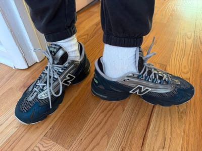 The image shows a person wearing New Balance 1000 sneakers, grey socks, and dark-colored sweatpants, standing on a wooden floor. The sneakers are primarily navy blue and silver with grey laces and have the New Balance "N" logo on the sides. The photo is taken from an overhead perspective, looking down at the person's feet. The location appears to be indoors, potentially a home, and the wooden floor is light brown. While the location is in the United States of America, it cannot be determined that the location is specifically in the city of Morton Grove.
