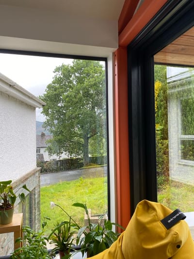 The image shows a view from inside a house located in Keswick, United Kingdom, at the coordinates 54.6 latitude and -3.12 longitude. The window frame is black and has an orange trim on the right side. The window is looking out at a tree, a street, and a house in the distance. There are several plants in the windowsill. A large, yellow beanbag chair sits in the bottom right corner of the image. The beanbag has a black strap attached to it with the word "extra" printed on it. It is raining outside, and the windows are wet.