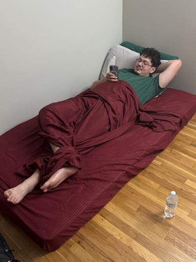 A slightly high-angle shot captures an indoor scene in Franklin Township, United States of America, showing a man lying on a bed or mattress on the floor, engrossed in his smartphone. The man, with dark curly hair, glasses, and a light beard, wears a dark green short-sleeved shirt. He lies on his left side, propped on a white pillow with a light gray pillowcase, with his right arm bent behind his head. His left hand holds a dark-colored smartphone, which has a distinct circular magnetic attachment visible on its back.
The bed is covered with a dark red sheet or blanket that features subtle striped patterns, and a large portion of it is draped over the man, covering his body with folds and wrinkles. His bare feet are visible peeking out from under the blanket at the lower right.
The setting includes a polished light brown hardwood floor with distinct plank patterns, running diagonally across the frame from bottom left to top right. A clear plastic water bottle with a white cap lies on the hardwood floor near the top right of the frame, casting a shadow. The background wall is a plain, light gray or off-white color. A dark bag is partially visible in the bottom right corner, next to the bed. The overall impression is one of relaxation or leisure. No discernible text is visible on any objects.