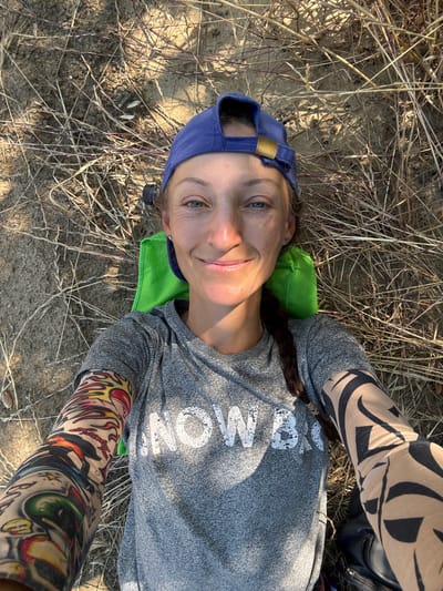A woman lies on the ground in a dry, grassy area. She is wearing a blue baseball cap, a grey t-shirt with the words "SNOW BIRD" printed on it, and has colorful tattoos on her arms. She is using a bright green object as a pillow. The ground is covered with dry grass and dirt. The lighting suggests it is daytime, possibly late afternoon due to the shadows cast by the sun. The location is the city of Gurzufsky Poselkovy Sovet, Ukraine.