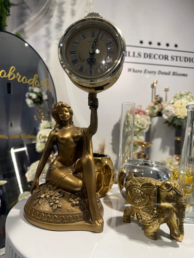 This image depicts a retail display in Ilidža, Bosnia and Herzegovina, showcasing various decorative items. The foreground features a golden figurine of a woman holding a clock aloft, seated on a decorative base. To the right of the figurine, a small golden elephant statue is visible, with intricate carvings. In the background, floral arrangements in vases, glass cylindrical vases, and a large black circular mirror with text are present. A sign with the text "MILLS DECOR STUDIO Where Every Detail Blooms" is also discernible in the background. The overall setting suggests an interior shop environment, likely a home decor store, with items displayed on a white surface.