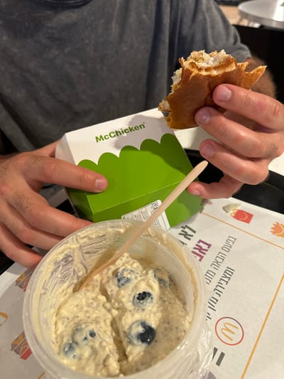 The image depicts a close-up of a person consuming food at a McDonald's restaurant, located in Airport, Israel. A person wearing a grey shirt holds a partially eaten McChicken sandwich in their right hand, while their left hand rests on the white and green 'McChicken' box. In the foreground, a clear plastic container contains a light-colored chia pudding or yogurt with visible blueberries, and a small wooden spoon is inserted. The food items are placed on a dark surface, overlaid with a McDonald's branded paper tray liner featuring Hebrew text and the red 'M' logo.