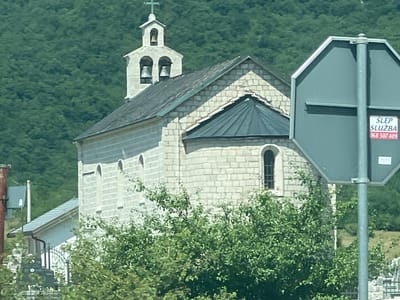 The image shows a stone church with a bell tower in the city of Dragalj, Montenegro. The church has a gray roof and a pointed arch window. A road sign is visible in the foreground, partially obscuring the church. The background is a lush green forest. The overall scene appears to be peaceful and rural.