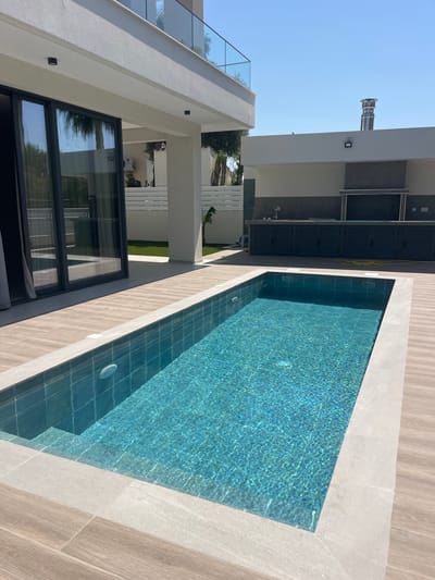 The image depicts a modern outdoor swimming pool area adjacent to a residential building. The pool itself is rectangular with turquoise water, tiled interior, and a light-colored coping. The surrounding deck is paved with light-brown wooden planks and the same light-colored stone used for the pool coping. To the left, a large glass sliding door opens from the building, reflecting palm trees and the sky. The building has a white facade with a balcony on the upper level, featuring a glass railing. To the right, an outdoor kitchen area is visible, with dark cabinets and a gray tiled backsplash, topped by a stainless-steel chimney. The environment is sunny with a clear blue sky. The location is the Moni Community, Cyprus. There are no people or animals visible in the image.