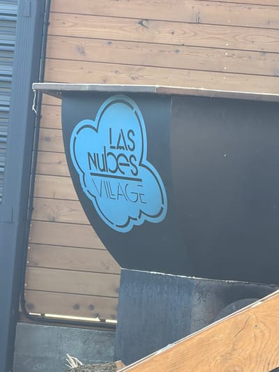 The image shows a close-up of a sign with a blue cloud outline and text in Russian. The text reads "LAS NUBES VILLAGE". The sign is mounted on a dark surface, possibly metal or painted wood, which is attached to a wooden wall. The wooden wall is made of horizontal planks. To the left of the sign is a vertical dark grey ribbed pipe or conduit. Below the sign is a concrete structure. In the lower right corner, a piece of angled wooden planking is visible. The scene appears to be outdoors. The sign is the primary subject of the image.