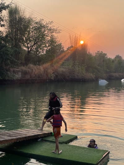 The image depicts a lakeside scene at dusk in Tambon Nam Phrae, Thailand. An orange sun hangs low in the sky, silhouetting a flock of birds and a tree with leafless branches. A wooden pier extends into the calm, reflective water. Two people stand at the end of the pier, one seemingly assisting the other to enter the water. In the water, a third person wearing goggles and a flotation device appears to be swimming. A small, white structure sits further back on the opposite shore, partially obscured by trees.