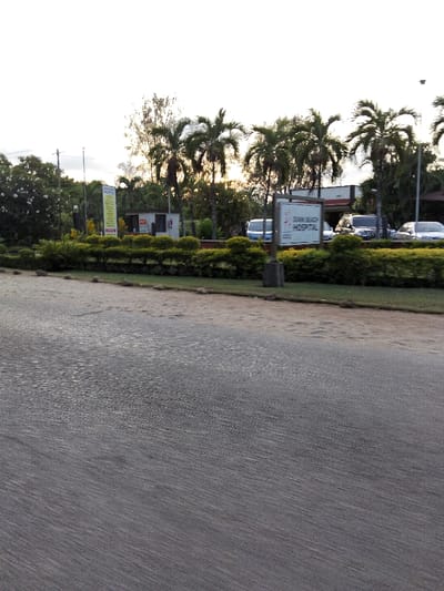 The image displays an outdoor scene in Gombato Bongwe ward, Kenya, featuring a paved road in the foreground. The road surface is dark gray with discernible texture. Adjacent to the road, a narrow strip of light-colored ground precedes a manicured green hedge. Beyond the hedge, an establishment is visible. On the left, a tall, vertical multi-panel sign stands amidst foliage. Centrally positioned, a white rectangular sign on a dark post clearly displays "DIANI BEACH HOSPITAL" with a logo below. Behind these signs, a small, light-colored building, possibly a security booth, is visible on the left, while portions of a larger building, consistent with a hospital entrance, are seen to the right. Several vehicles are present within the establishment's area: a light-colored van or minibus is visible to the left of the hospital sign, and multiple dark-colored SUVs or cars are parked or positioned further right. The background is characterized by numerous tall palm trees and dense green foliage under a light, hazy sky. No individuals are visible in the scene.