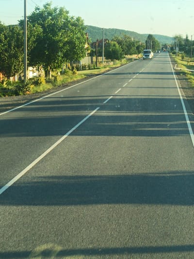 The image shows a two-lane asphalt road in the vicinity of Nevytske, Ukraine. The road is lined with white dashed and solid lines. To the left of the road, there are trees, houses, and a utility pole. To the right, the road is also bordered by trees and vegetation. In the distance, rolling hills covered in trees are visible under a clear sky. A white van is driving on the road, heading away from the viewer. The scene appears to be captured during daylight, possibly in the late afternoon, given the long shadows cast across the road.