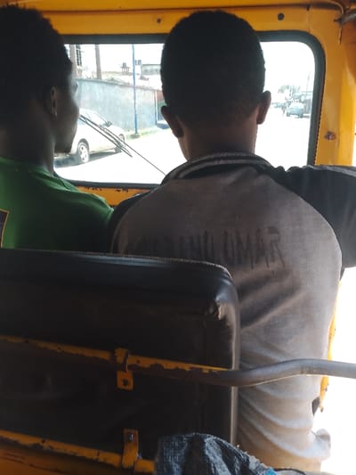 Inside a yellow auto rickshaw, two individuals are seated. One wears a green shirt, facing towards the street visible through the open window, where a white car is parked. The other, wearing a grey shirt, faces the camera, with the name "KUNO UMAR" printed on the back. A dark, worn-out seat is in the foreground. The rickshaw appears to be moving along a street, indicated by the visible vehicles and buildings outside.