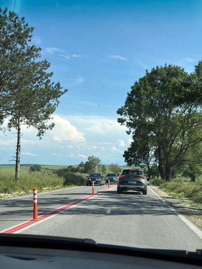 The image, taken in Brestovets, Bulgaria, captures a bright, sunny scene from inside a vehicle, looking forward along a rural road. The perspective shows part of the vehicle's dashboard or interior on the far right.
A line of cars is visible on the asphalt road, seemingly queuing or moving slowly. The most prominent vehicle is a dark green Kia SUV, seen from the rear, displaying the "KIA" logo and a Bulgarian license plate: "DB 0404 BB". Behind it, a black car is partially visible. The road features a distinct solid red line running along its right side, with orange and white traffic delineators (bollards) placed intermittently on this line, suggesting a temporary lane division or guidance.
The surrounding landscape is verdant and open. To the left of the road, large, leafy green trees cast long, sharp shadows across the asphalt, indicating bright sunlight, likely mid-day or early afternoon. Further to the left, a tall pine tree with a slender, somewhat barren trunk stands out against the sky. Beyond the trees and road, expansive green fields stretch into the distance under a vast, clear blue sky adorned with scattered white cumulus clouds.
The overall scene conveys a calm moment of travel through a sunny, picturesque Bulgarian countryside. No people are visible in the frame.