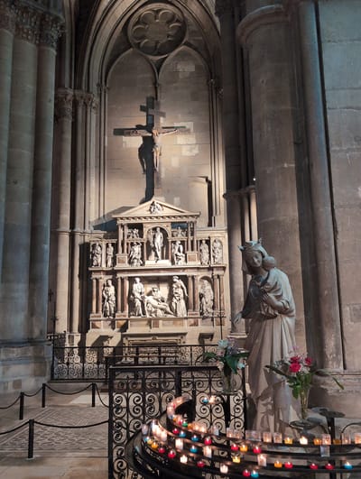 The photograph depicts the interior of a stone church in Urvillers, France. In the center of the frame, a large crucifix with a figure of Christ is mounted on a stone wall above an ornate altar. The altar is decorated with multiple stone reliefs depicting biblical scenes and figures. To the right of the altar, a life-sized statue of a crowned woman holding a baby stands, with flowers placed in vases on either side. In the foreground, a black metal railing encloses a circular arrangement of lit votive candles of various colors. The architecture features tall, fluted stone columns, arched doorways, and stone walls. The lighting is dim, with spotlights illuminating the altar and crucifix, and the candles providing a warm glow in the foreground.