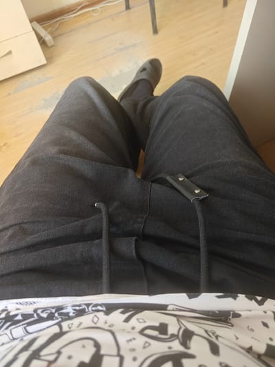 This media file shows a close-up view of a person's legs and lap area, seated on a wooden floor. The person is wearing dark wash denim jeans and black slides. The jeans have a drawstring detail at the waist, with a black leather tag secured by two rivets. A black cord extends from the drawstring area. The background shows a portion of a white cabinet or piece of furniture to the left, and a lighter colored wall with electrical wires along the baseboard. To the right, a vertical white surface is visible, possibly part of a wall or furniture. The overall scene suggests an indoor environment with simple furnishings. The lighting appears to be natural, coming from a window or overhead source, indicating it could be daytime. No specific weather conditions can be inferred. The location is identified as Dünggilagaş, Kazakhstan. The image focuses on the lower body of the person and the texture of their clothing and surroundings, creating a grounded and personal perspective. There is no visible text in the image.